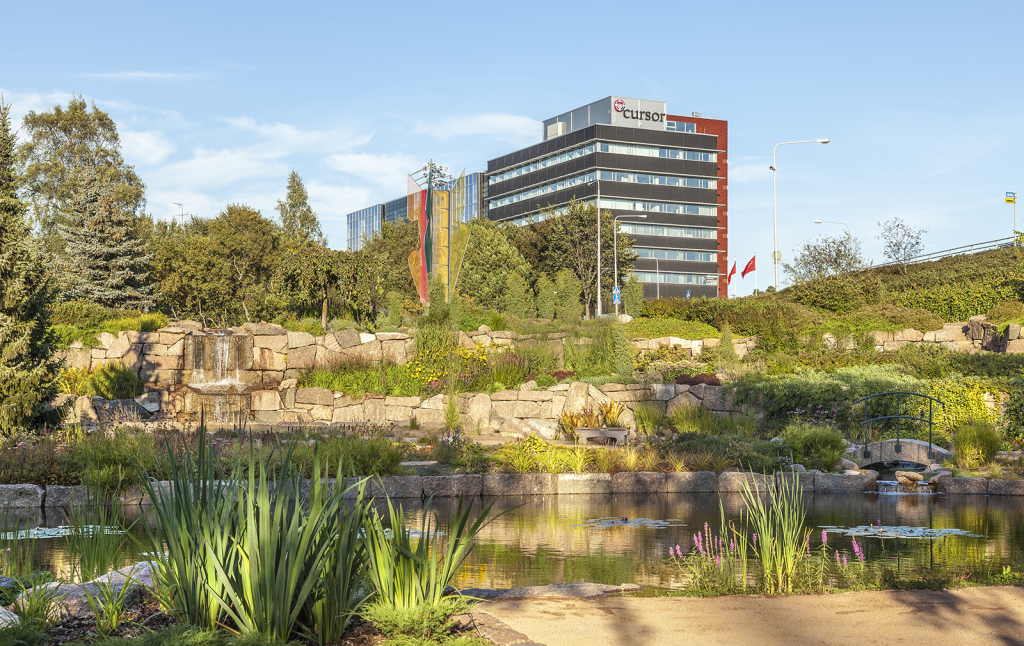 Karhulan jokipuiston maisemaa, jossa paljon vihreitä kasveja ja kaukana taustalla näkyy korkea toimistorakennus.